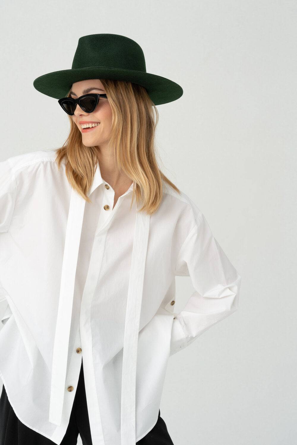 Woman wearing a dark green fedora hat with a white button-up shirt and black pants, exuding effortless elegance and timeless style.