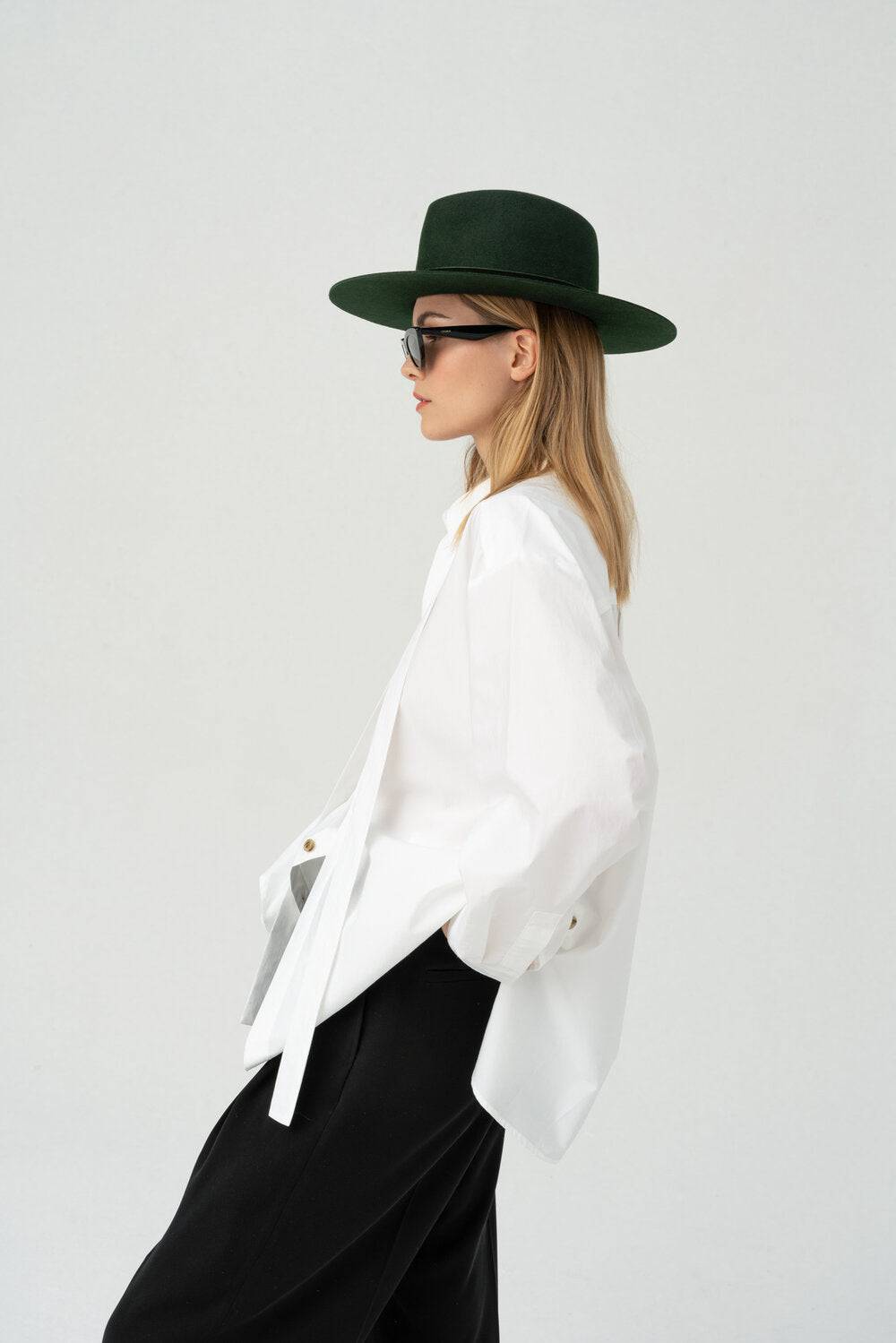 Side profile of a woman wearing a dark green fedora hat, paired with a white oversized shirt and black trousers for a modern, stylish outfit.
