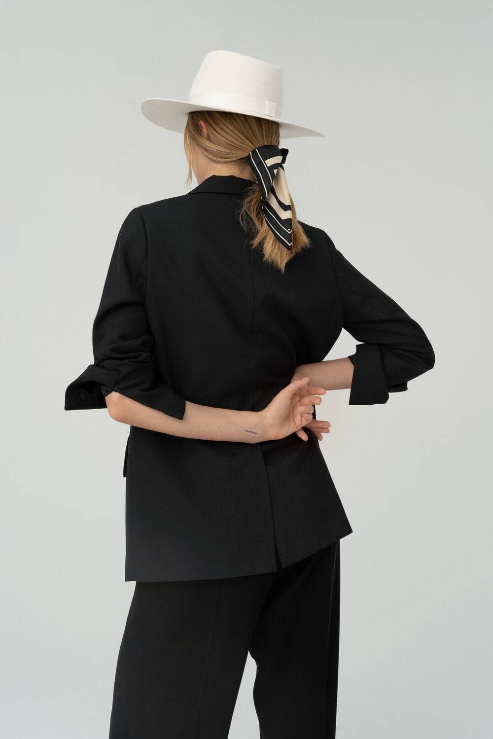 Back view of a woman wearing an ivory fedora hat with a monogram, complemented by a black blazer and a stylish ribbon accent.