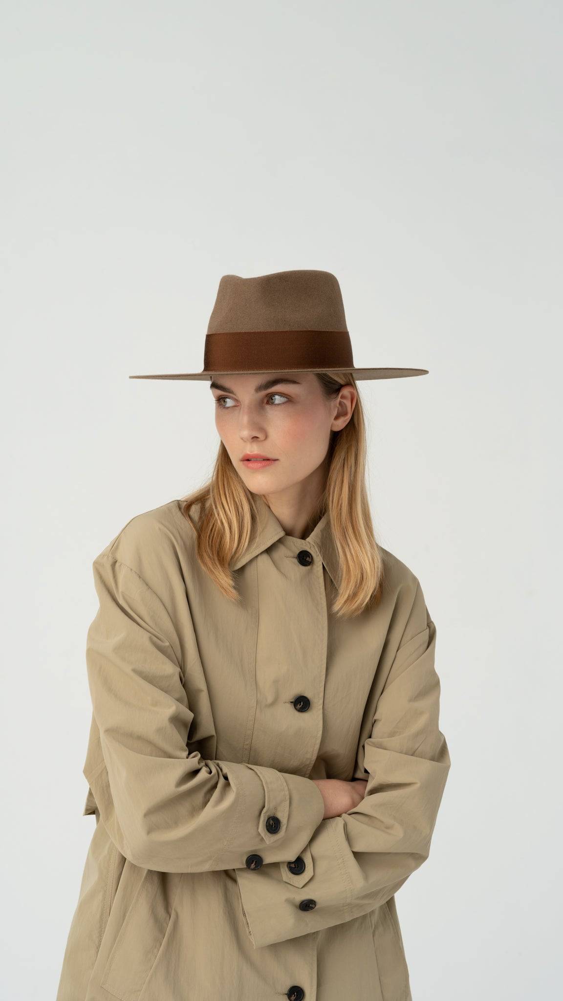 Close-up of a model in a walnut fedora hat with a brown band, styled with a beige trench coat. A handcrafted, dress-weight hat for a refined look.