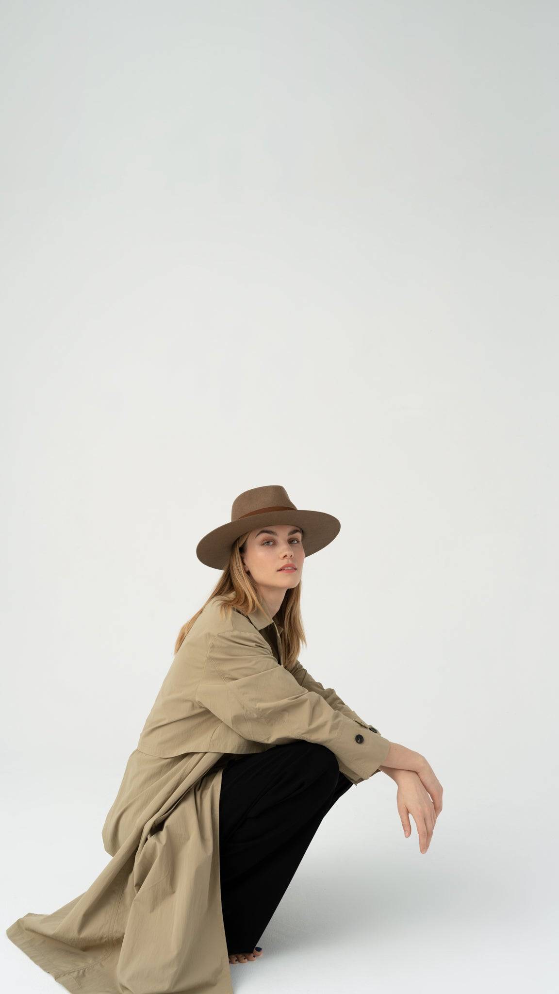 Model wearing a walnut fedora hat with a brown band, crouching in a stylish beige trench coat. A perfect autumn accessory with timeless appeal.