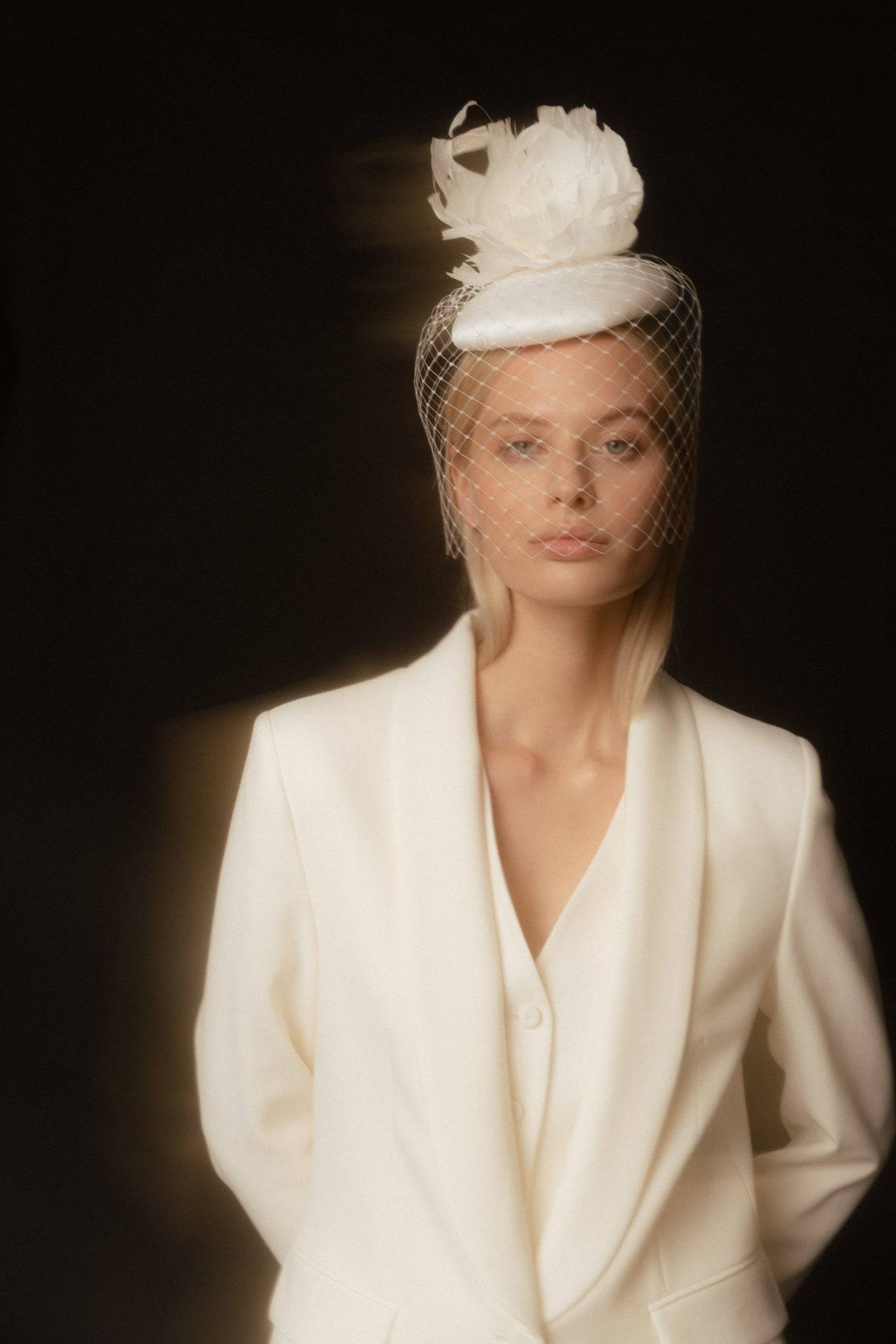 Luxury bridal cocktail hat adorned with a feather flower and birdcage veil. Handcrafted white silk wedding headpiece for a sophisticated, vintage look.
