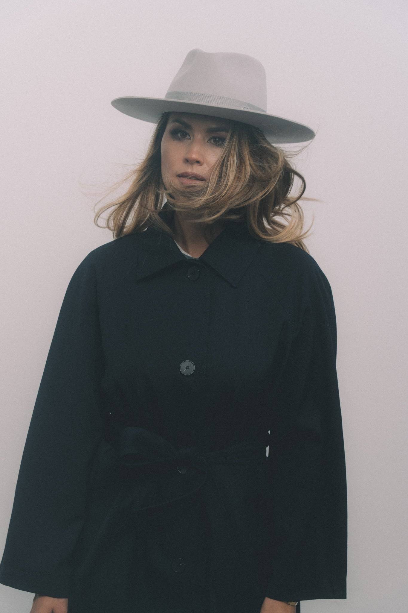 Light grey fedora hat with monogram embellishment, styled with a black trench coat. Crafted from rabbit fur for a timeless, elegant look.