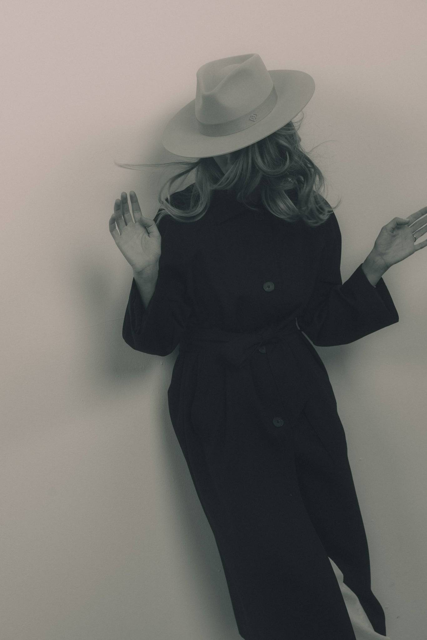 Artisan light grey fedora hat with a monogram detail, paired with a black coat. A handcrafted designer piece for a refined look.
