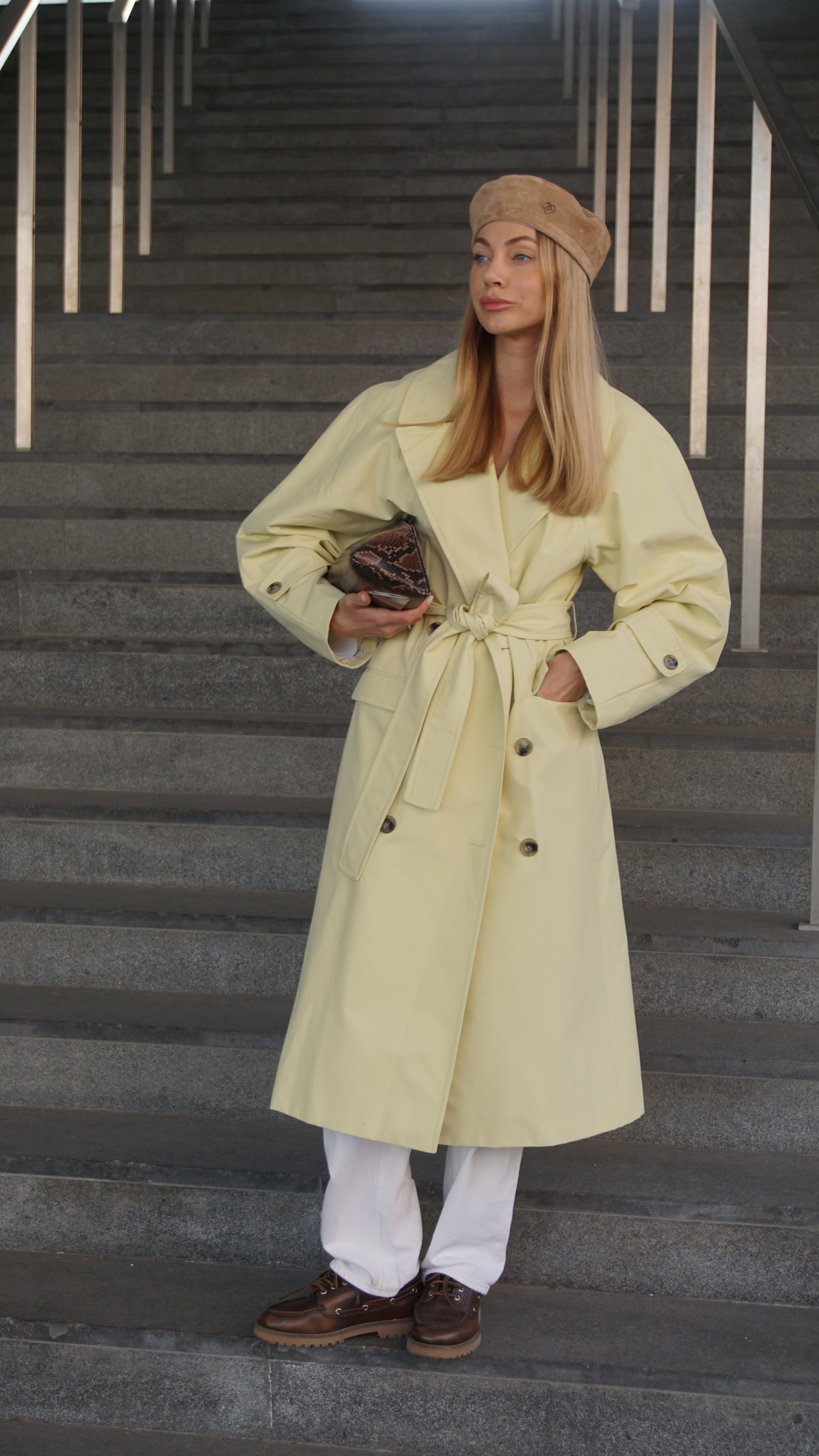 Woman wearing a light brown suede leather beret paired with a beige trench coat, white pants, and brown loafers. Stylish Parisian-inspired outfit on urban stairs.