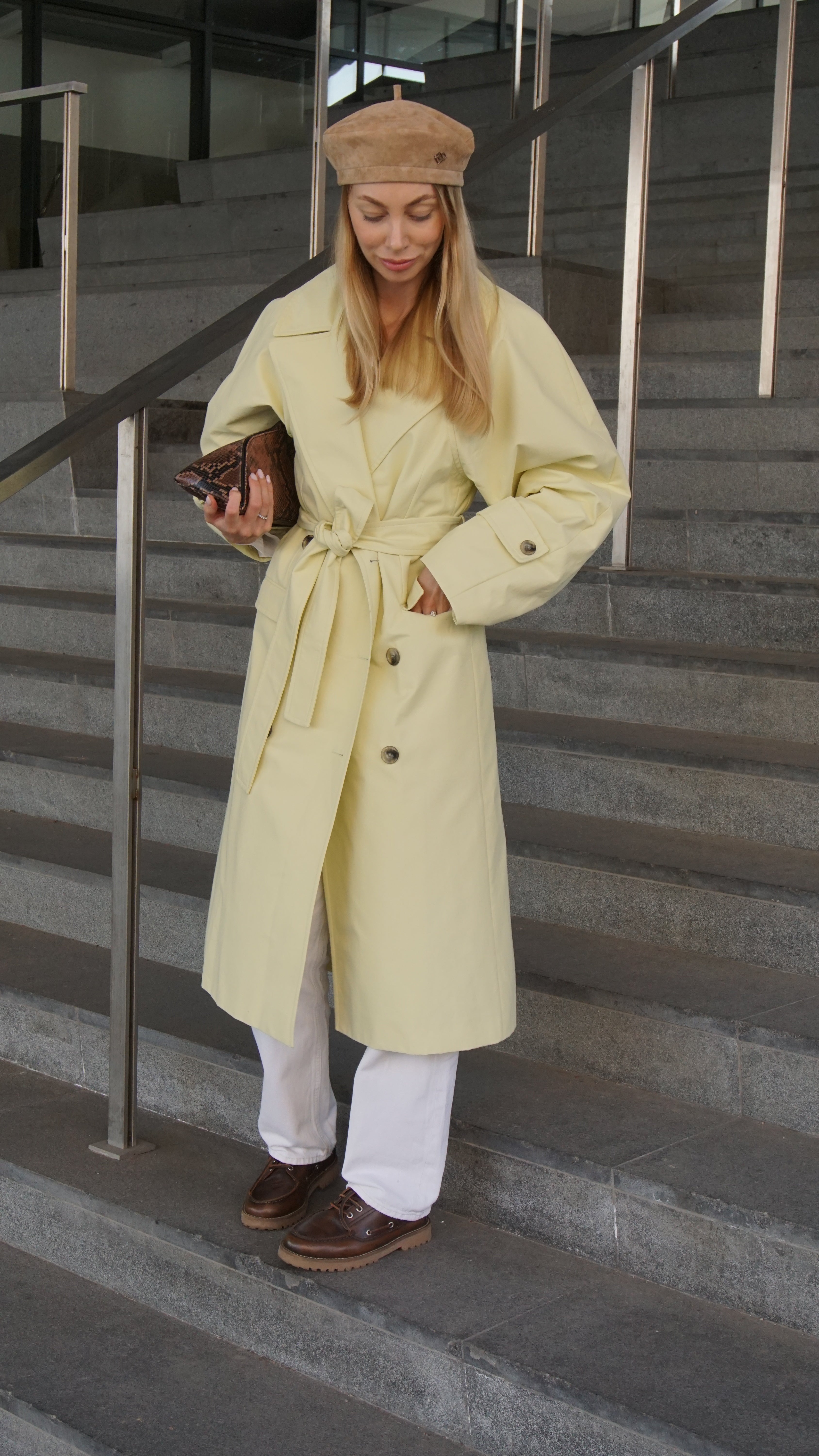 Woman wearing a light brown suede leather beret paired with a yellow trench coat and white pants. Stylish Parisian-inspired outfit with a classic beret.