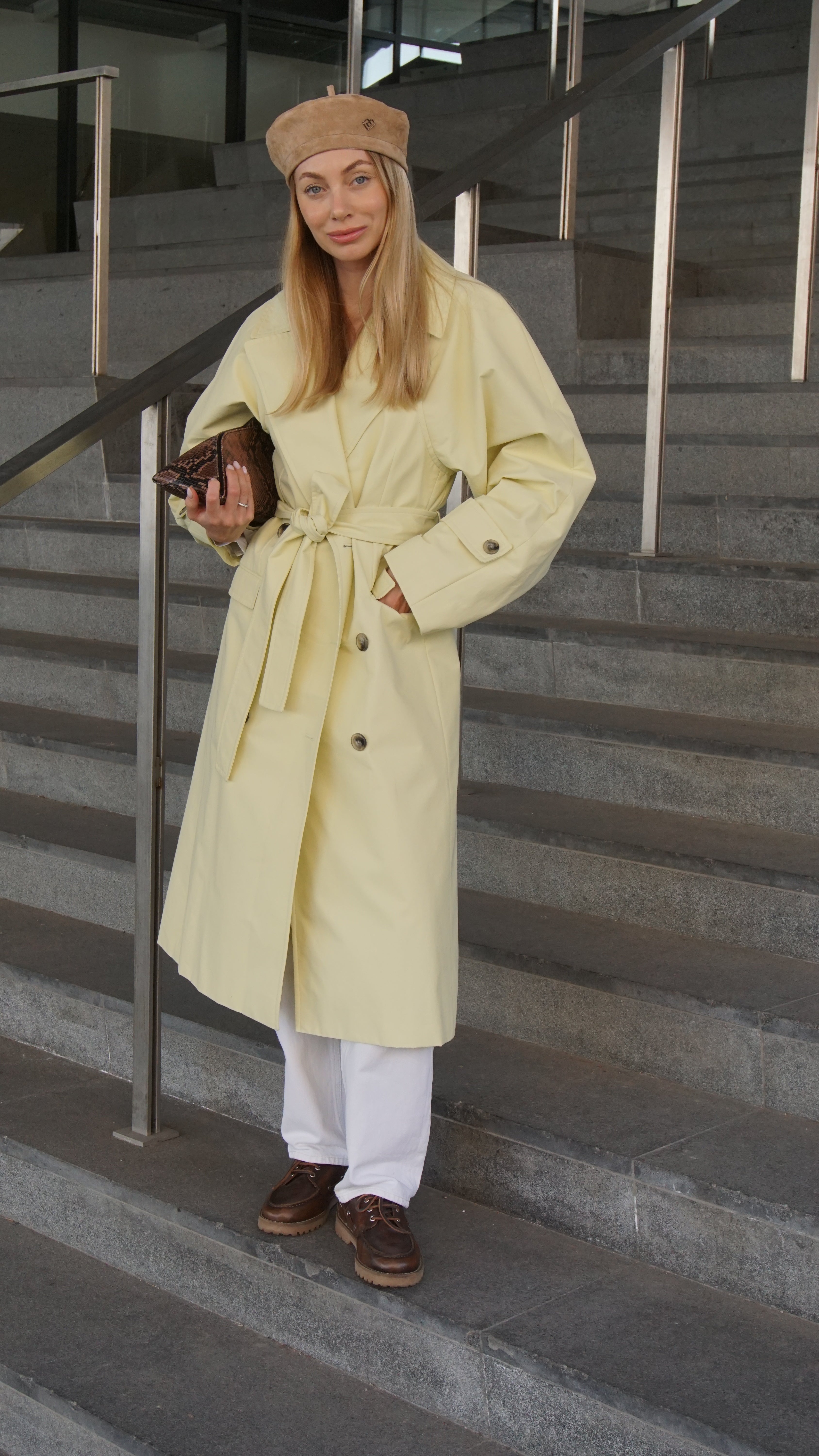 Woman wearing a light brown suede leather beret paired with a beige trench coat, white pants, and brown shoes. Stylish and modern outfit for a chic look.