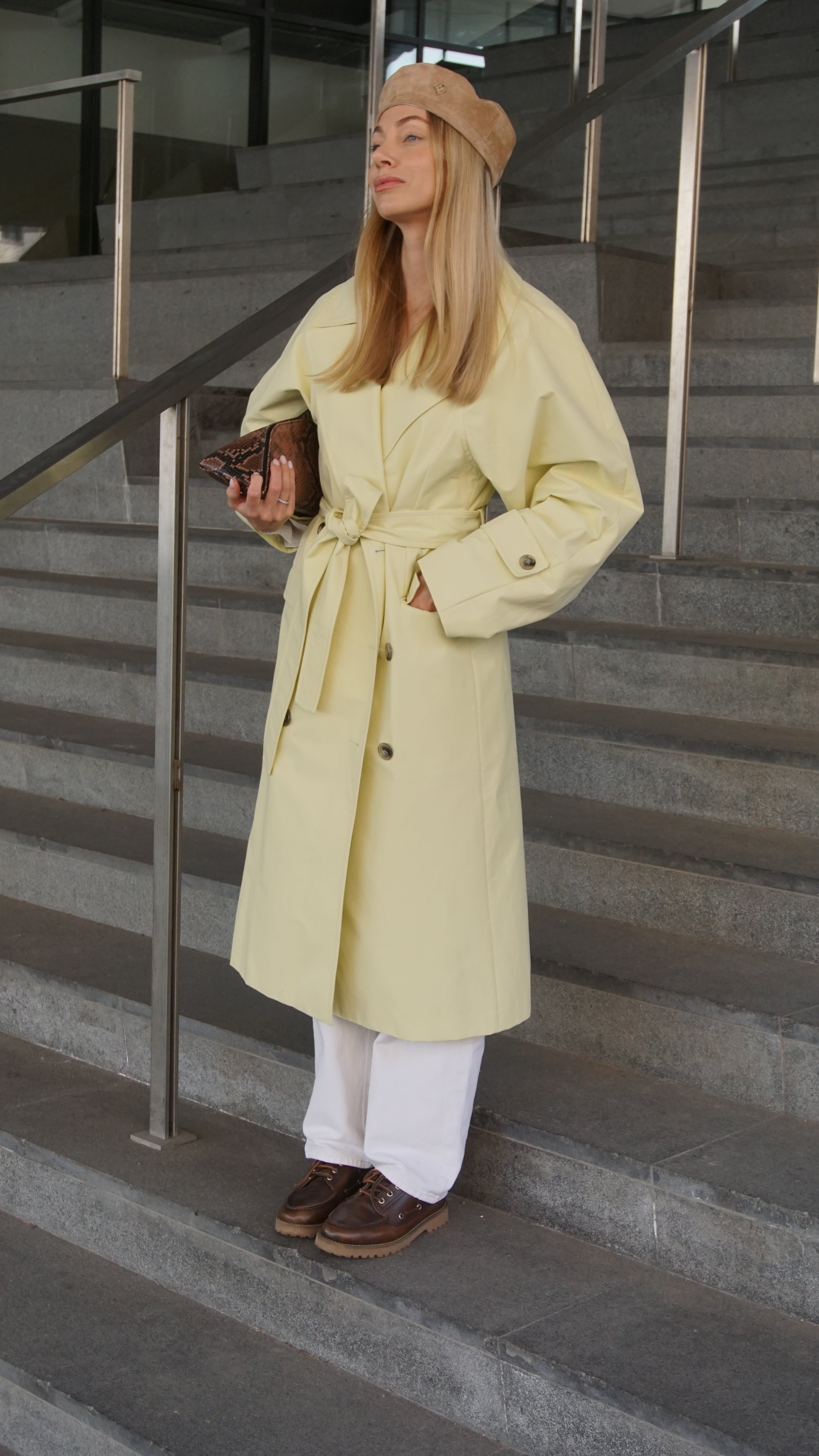 Woman wearing a light brown suede leather beret paired with a yellow trench coat, white pants, and brown shoes, standing on stairs with a clutch in hand.