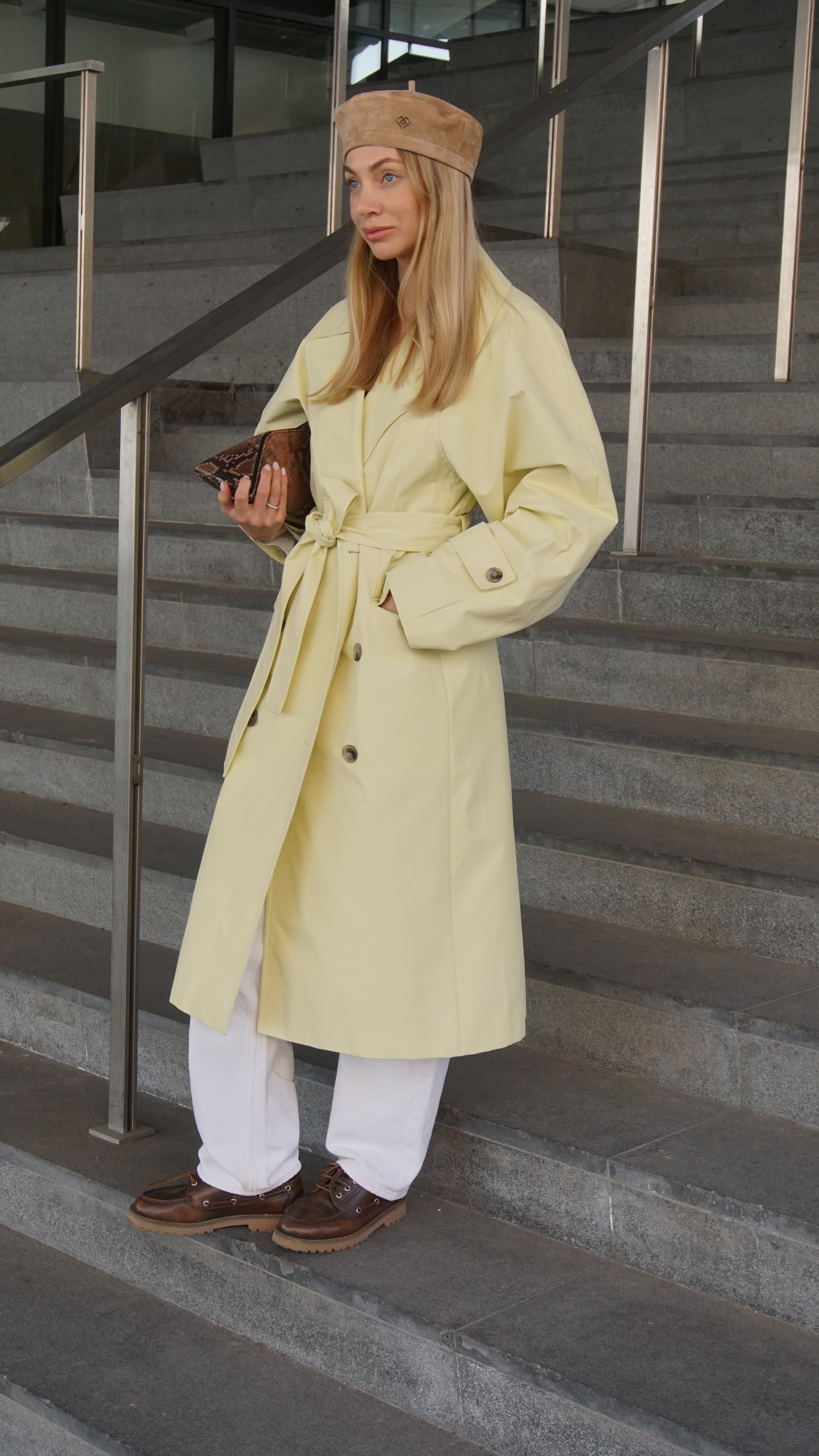 Woman wearing a light brown suede leather beret paired with a beige trench coat, white pants, and brown shoes. Stylish streetwear fashion with modern accessories.