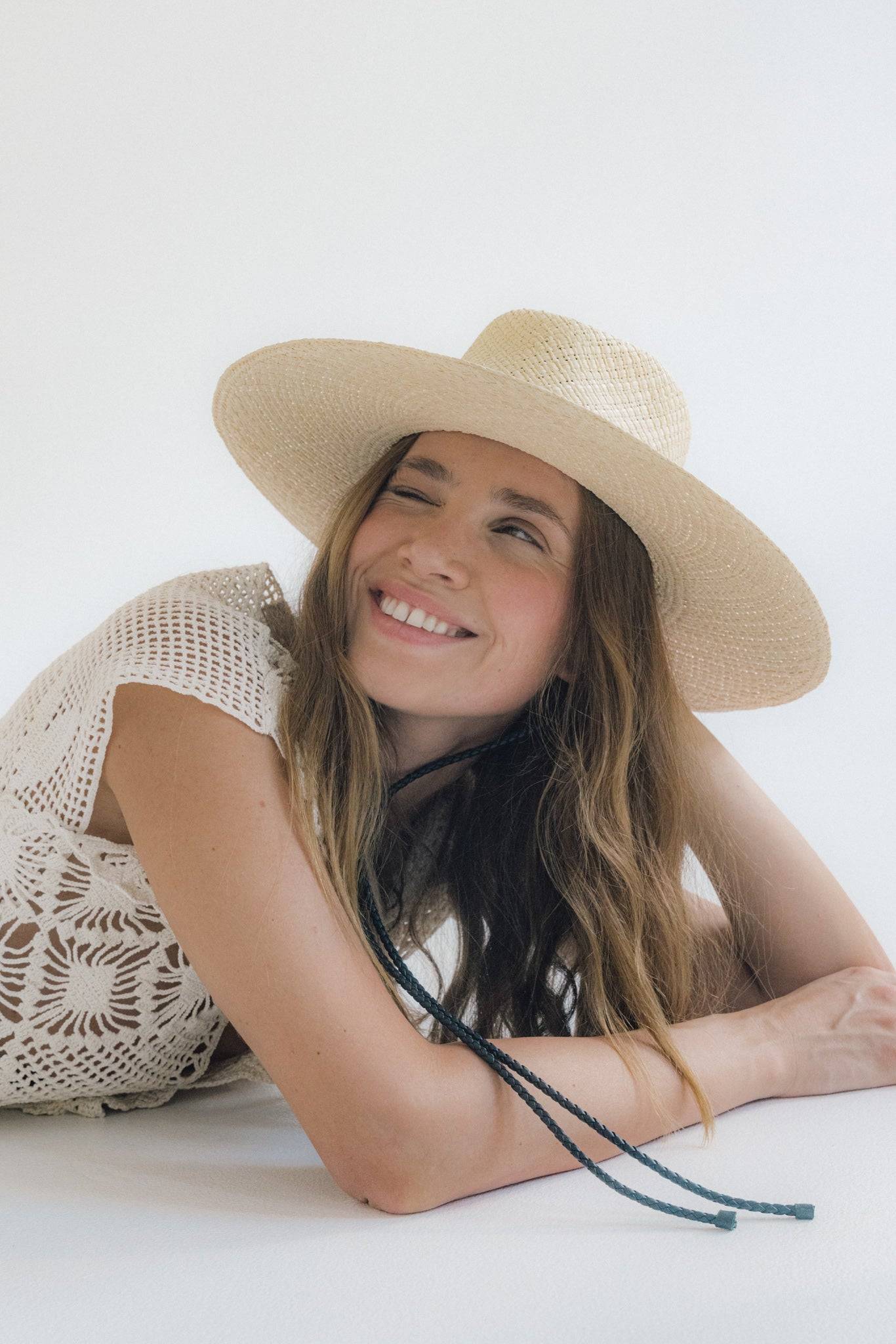 Smiling woman wearing a straw fedora hat with a leather strap, lying down in a relaxed pose. A must-have summer accessory for effortless style.