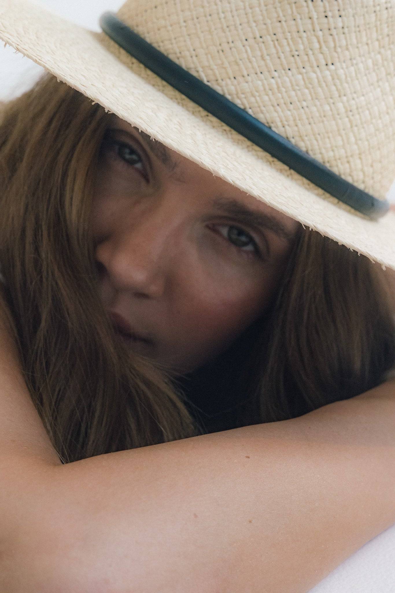 Close-up of a woman partially hiding behind her arm, showcasing the texture of a straw fedora with a leather strap. A timeless summer essential.