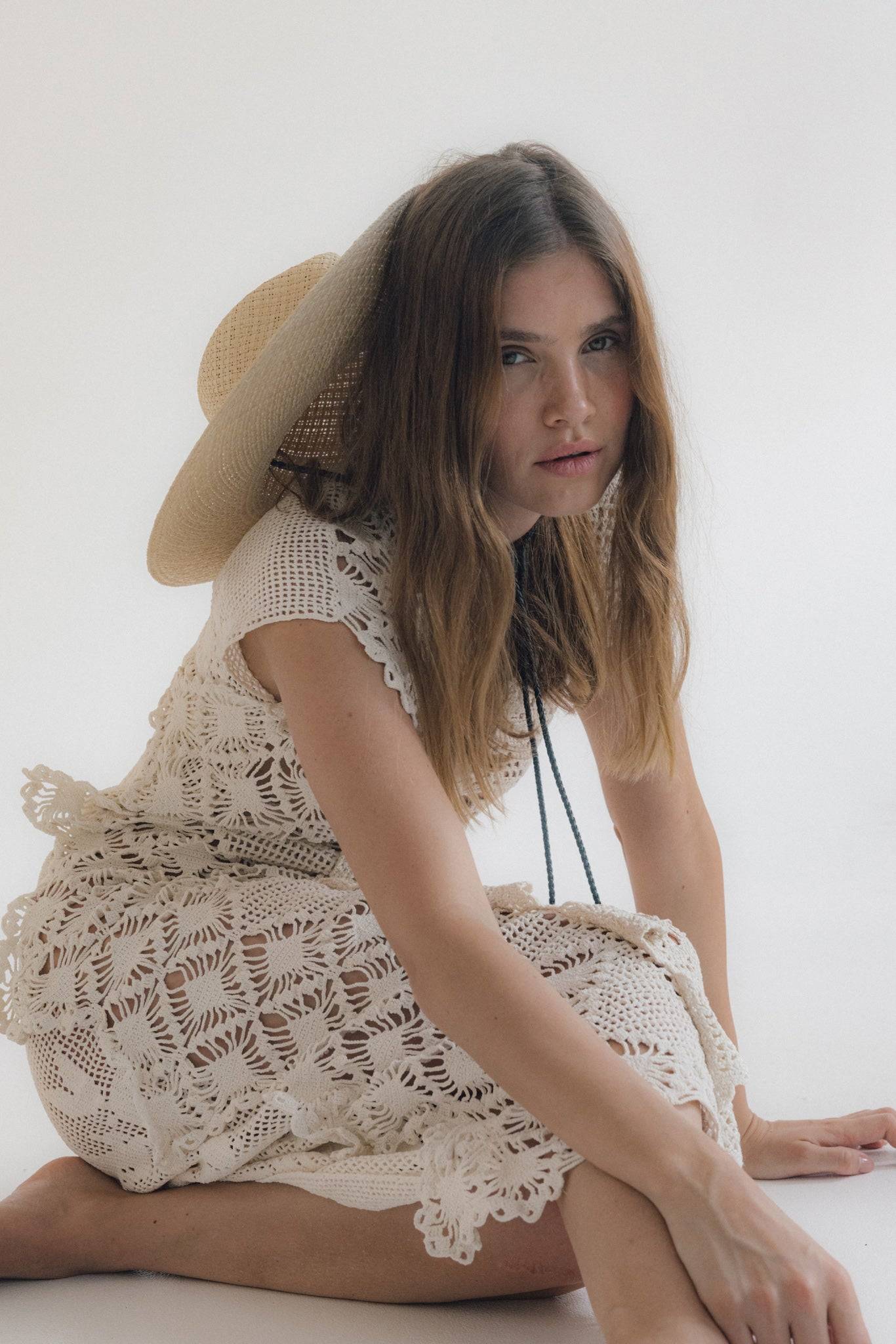 Model crouching in a lace dress with a straw fedora hat resting on her back. A chic and breathable summer outfit for warm weather styling.