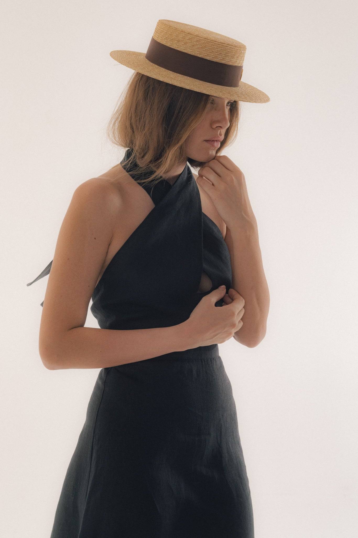 Fashionable woman in a black dress adjusting her monogram-embellished straw boater hat, ideal for a refined summer look.