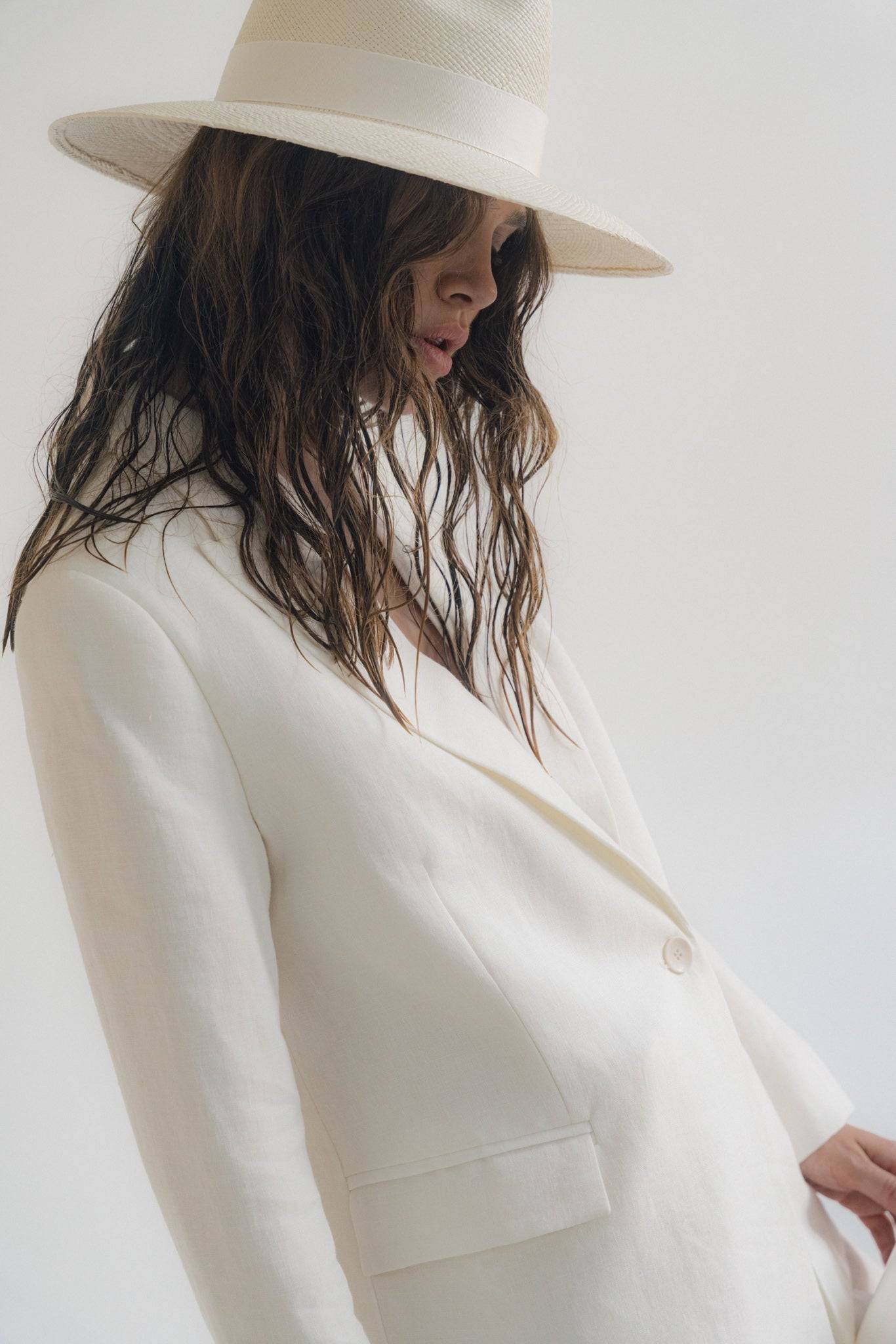 Side profile of a model wearing a white wide-brimmed fedora hat with a structured white blazer. A statement piece for a sophisticated summer look.