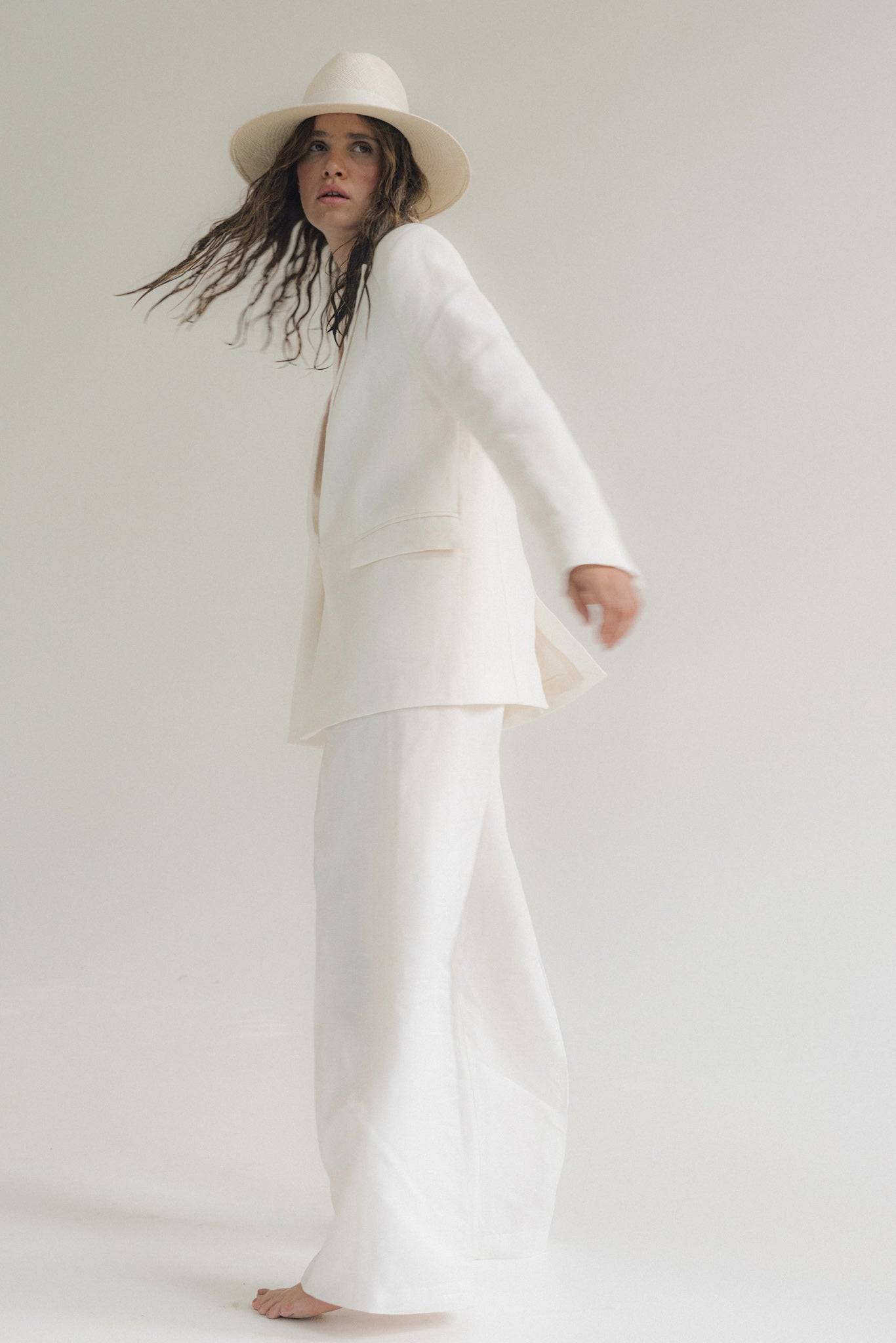 Dynamic shot of a model in motion wearing a white wide-brimmed fedora hat and a flowing white suit. A chic summer accessory for modern elegance.