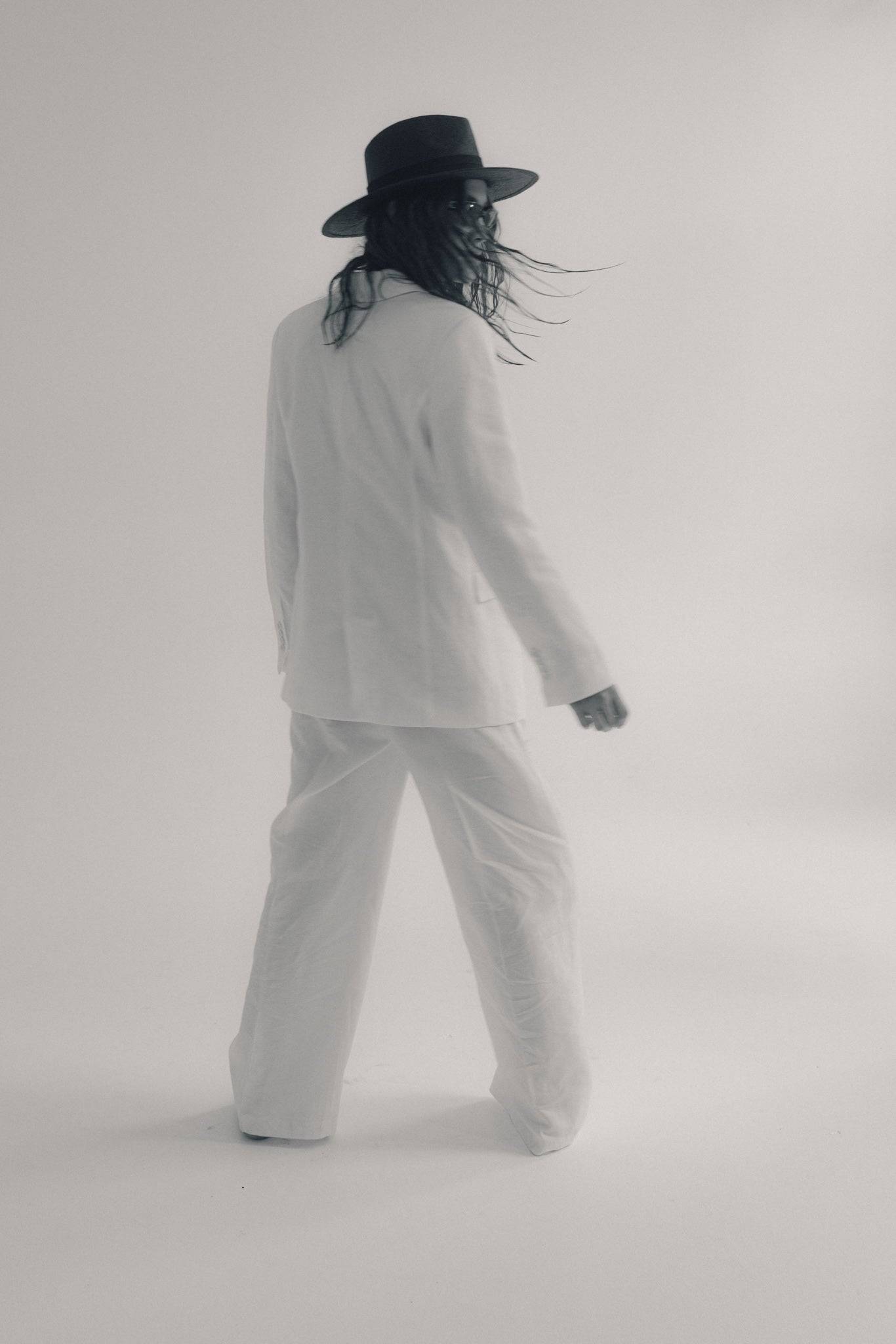 Back view of a model in a black fedora straw hat and white suit, showcasing a breezy, sophisticated summer style with movement.