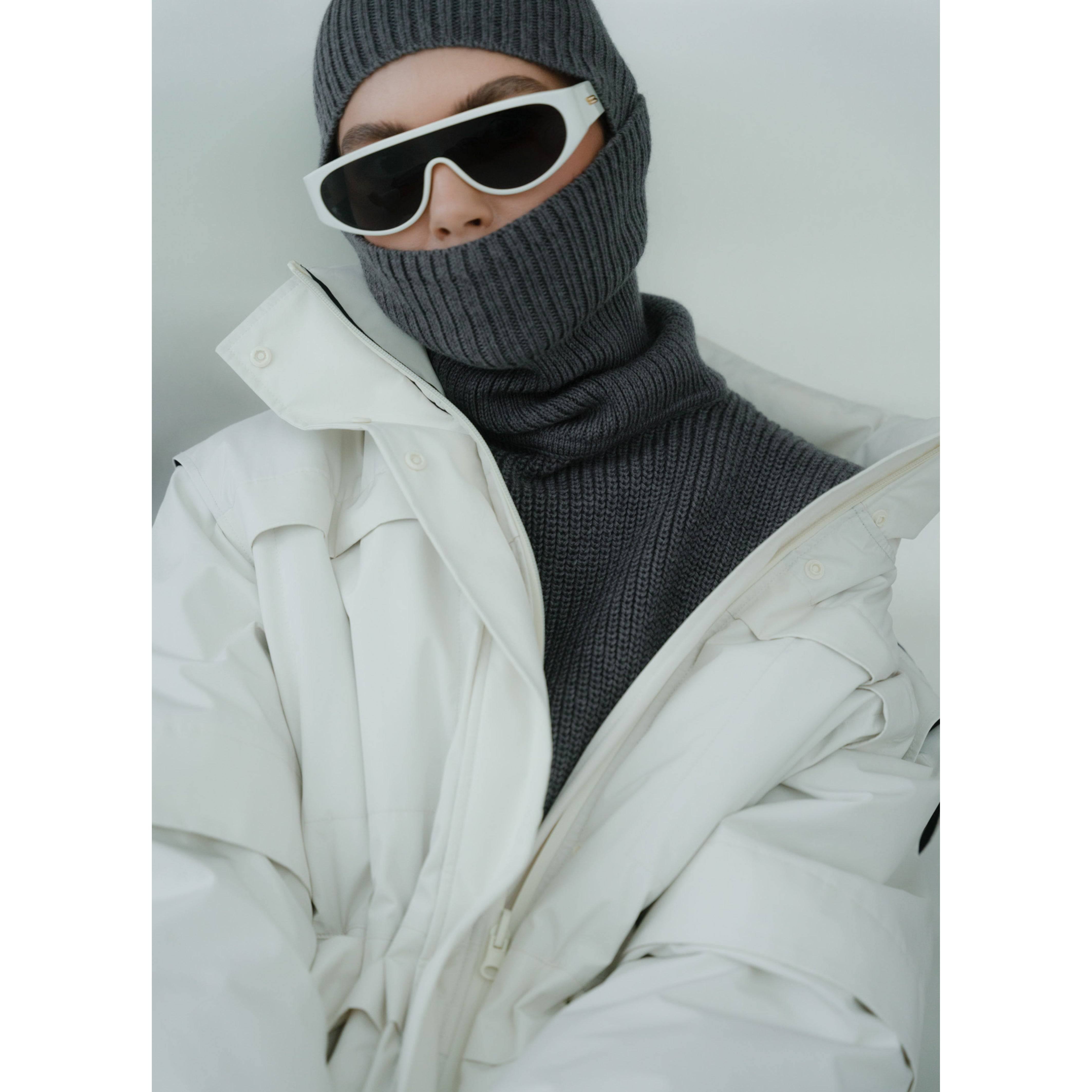Close-up of a dark grey Merino wool balaclava paired with a white winter coat and sunglasses. A chic and functional cold-weather accessory.