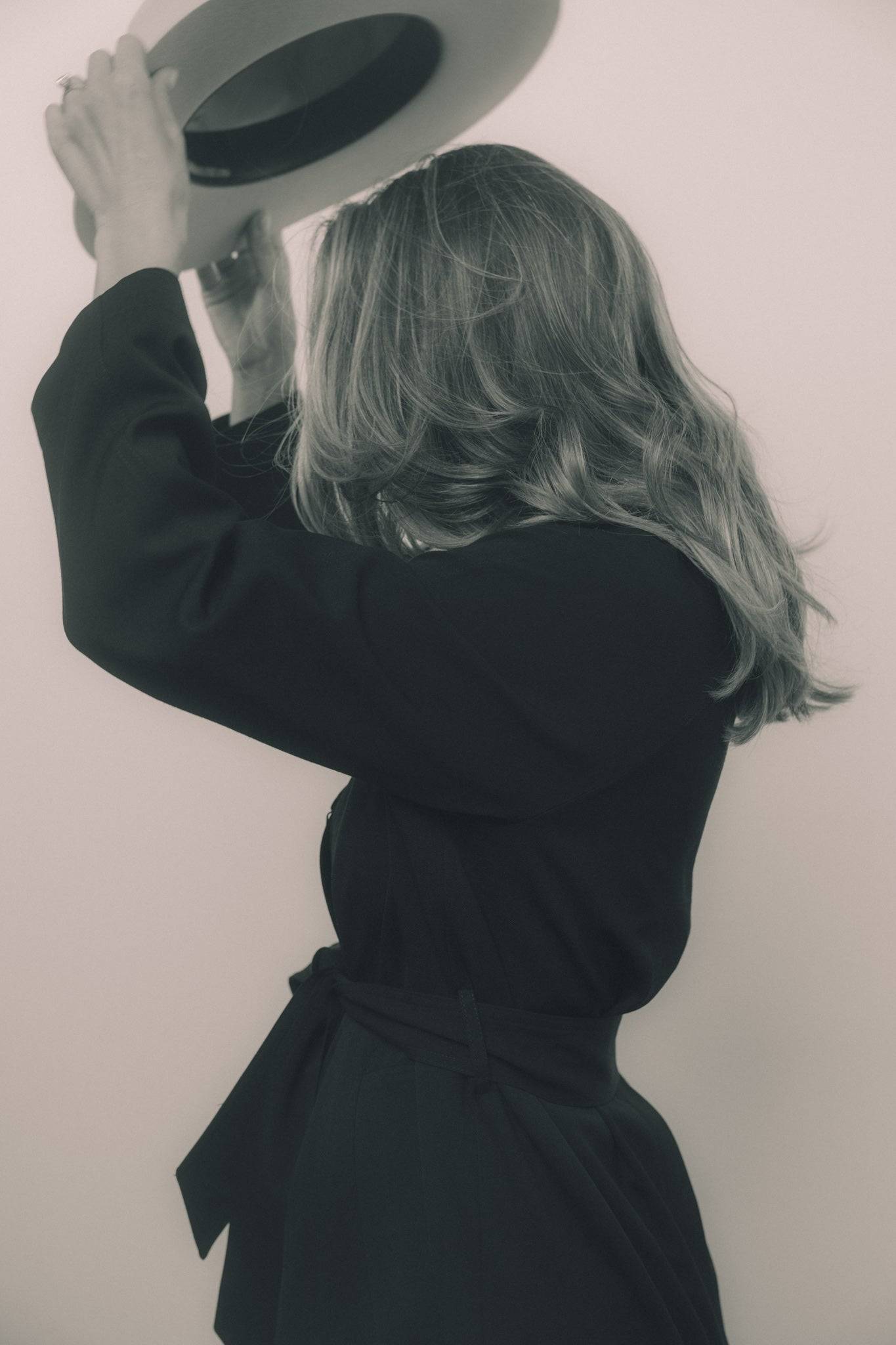 Monochrome image of a woman lifting an ivory fedora hat, showcasing its handcrafted quality and sophisticated design.