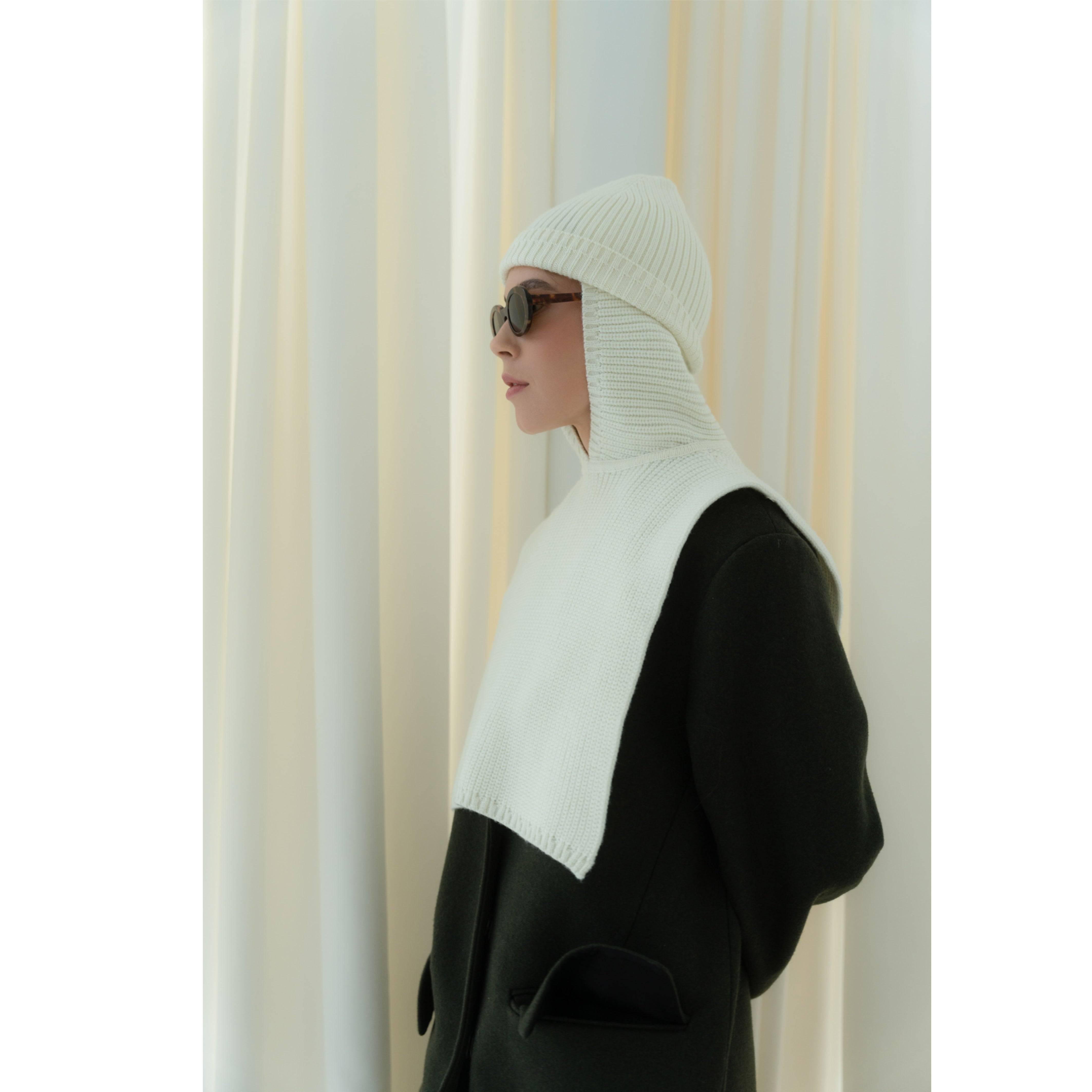 Side view of a model in a milky white Merino wool balaclava, showcasing its ribbed knit texture and elegant drape over a dark coat.